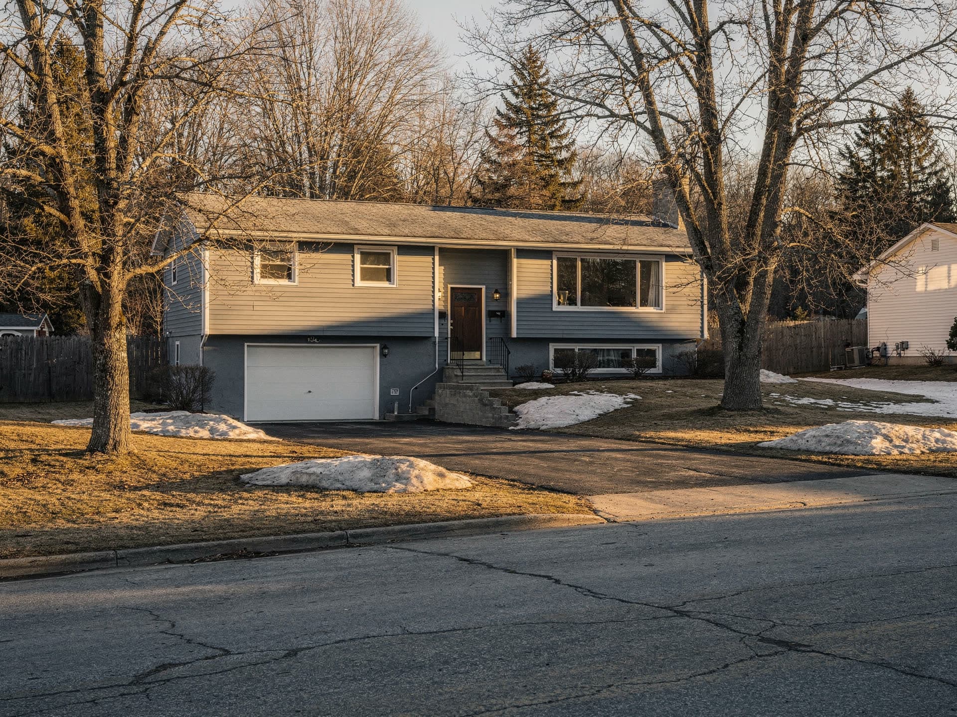 A blue grey split level house seen from across the street in low winter sun, bare trees and patches of melting snow on the lawn, the garage tucked in under the front door steps