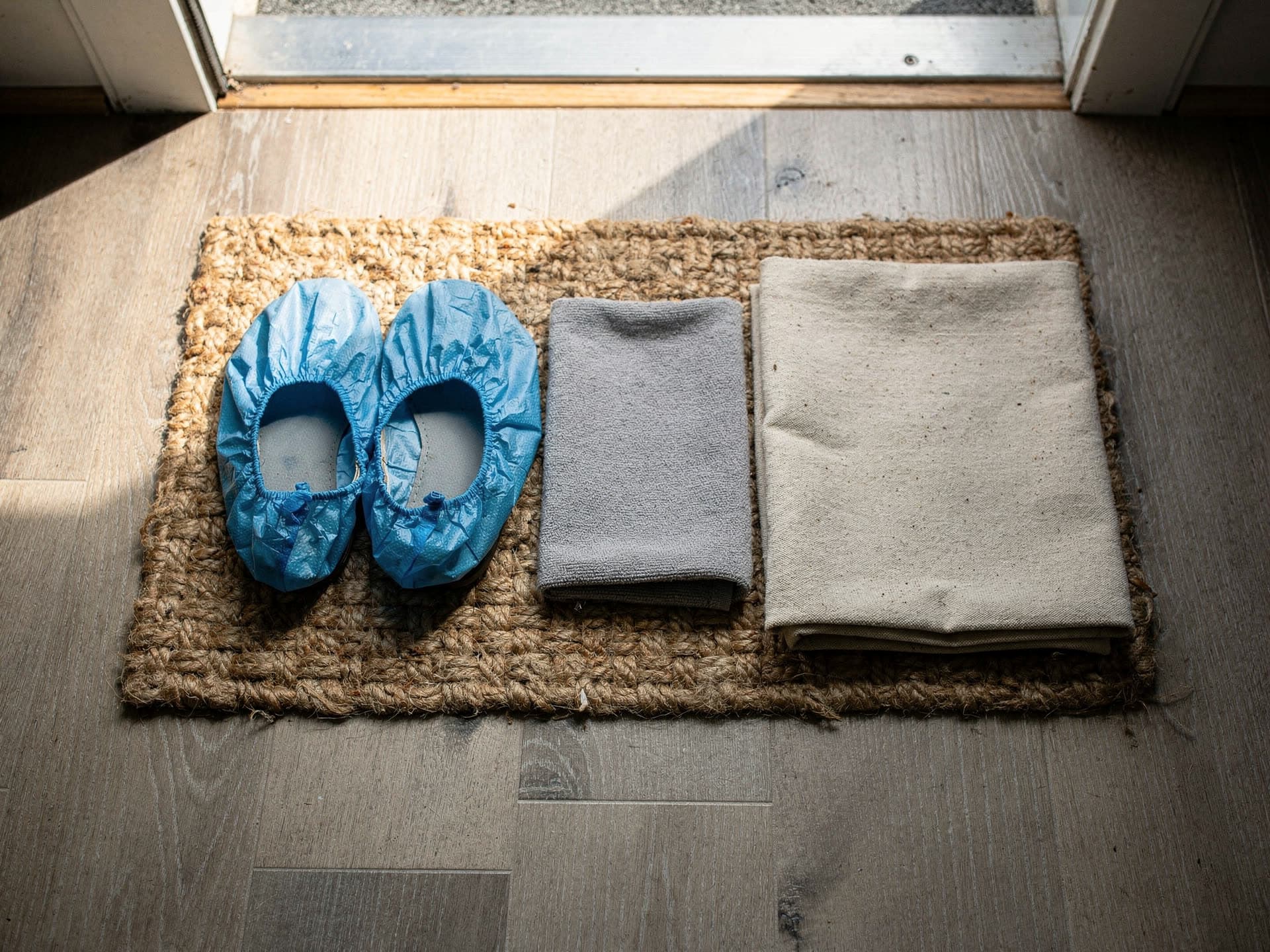 Looking straight down at a jute doormat inside a front door, holding a used pair of blue disposable shoe covers, a folded grey microfiber cloth and a folded square of natural canvas drop cloth, lit by daylight from the open door.