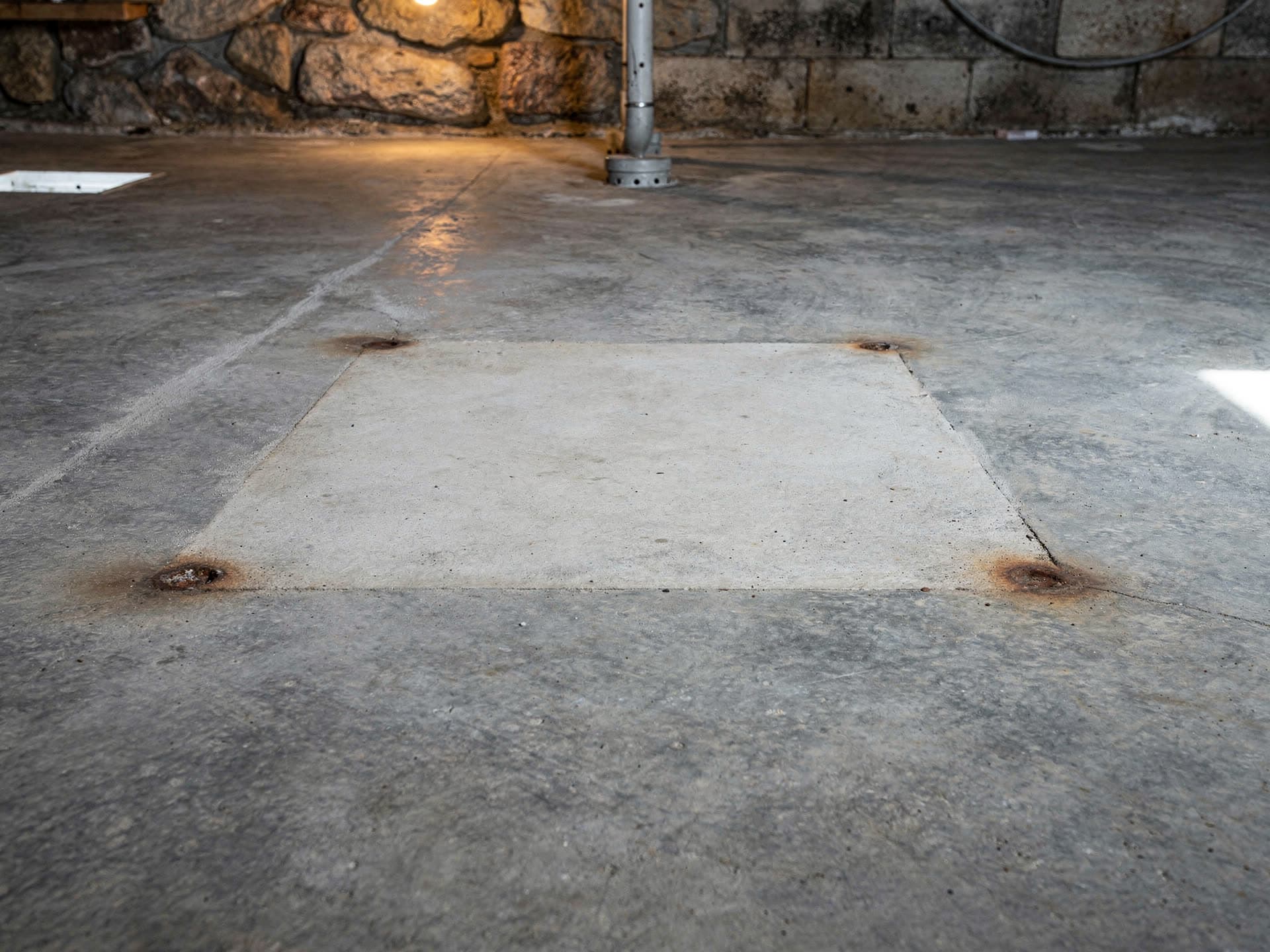 A swept grey concrete basement floor photographed from close to the ground, showing the clean pale rectangle and four rust rings left where a large old unit stood for years, with a stone and block foundation wall and a warm bulb behind.