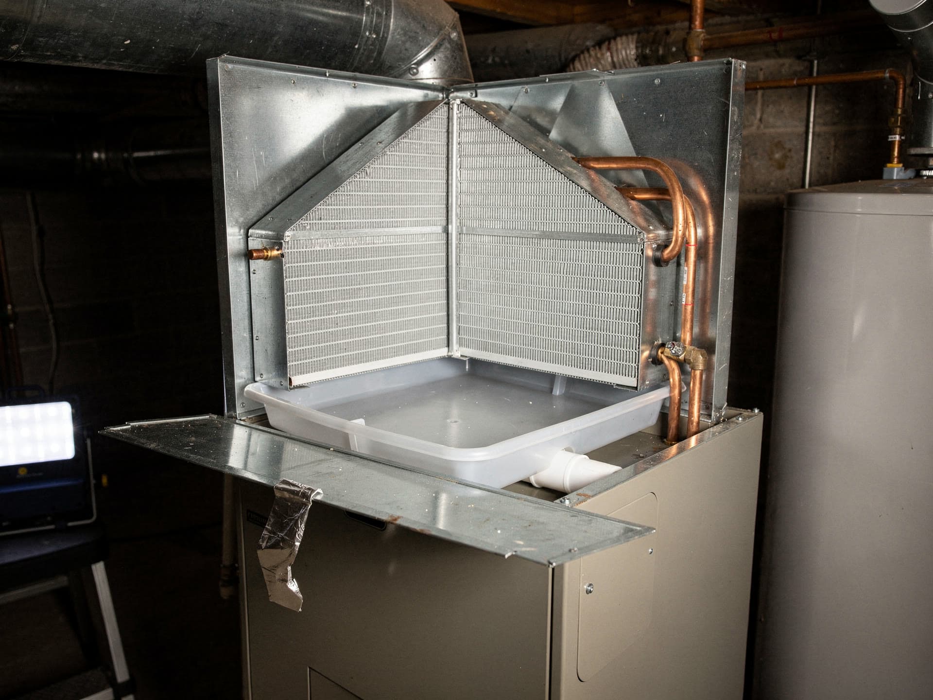 An A shaped evaporator coil sitting open on top of a furnace cabinet with the access panel hinged down, copper line set and brass fittings at its right side and the white condensate pan beneath it