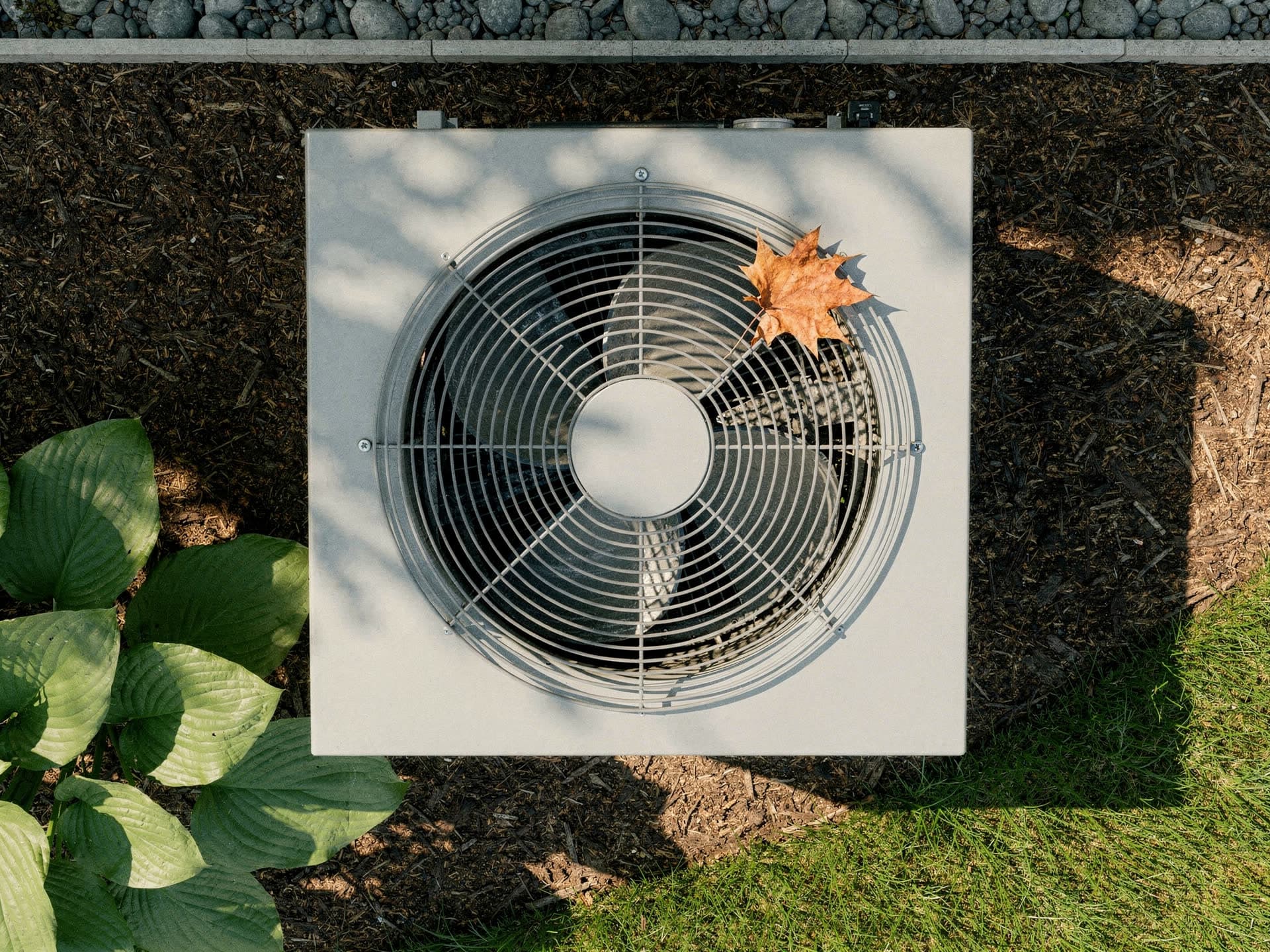 Looking straight down onto the fan grille of an outdoor condenser set in a mulch bed, a single fallen maple leaf resting on the guard, hostas at one edge of the bed and lawn at the other