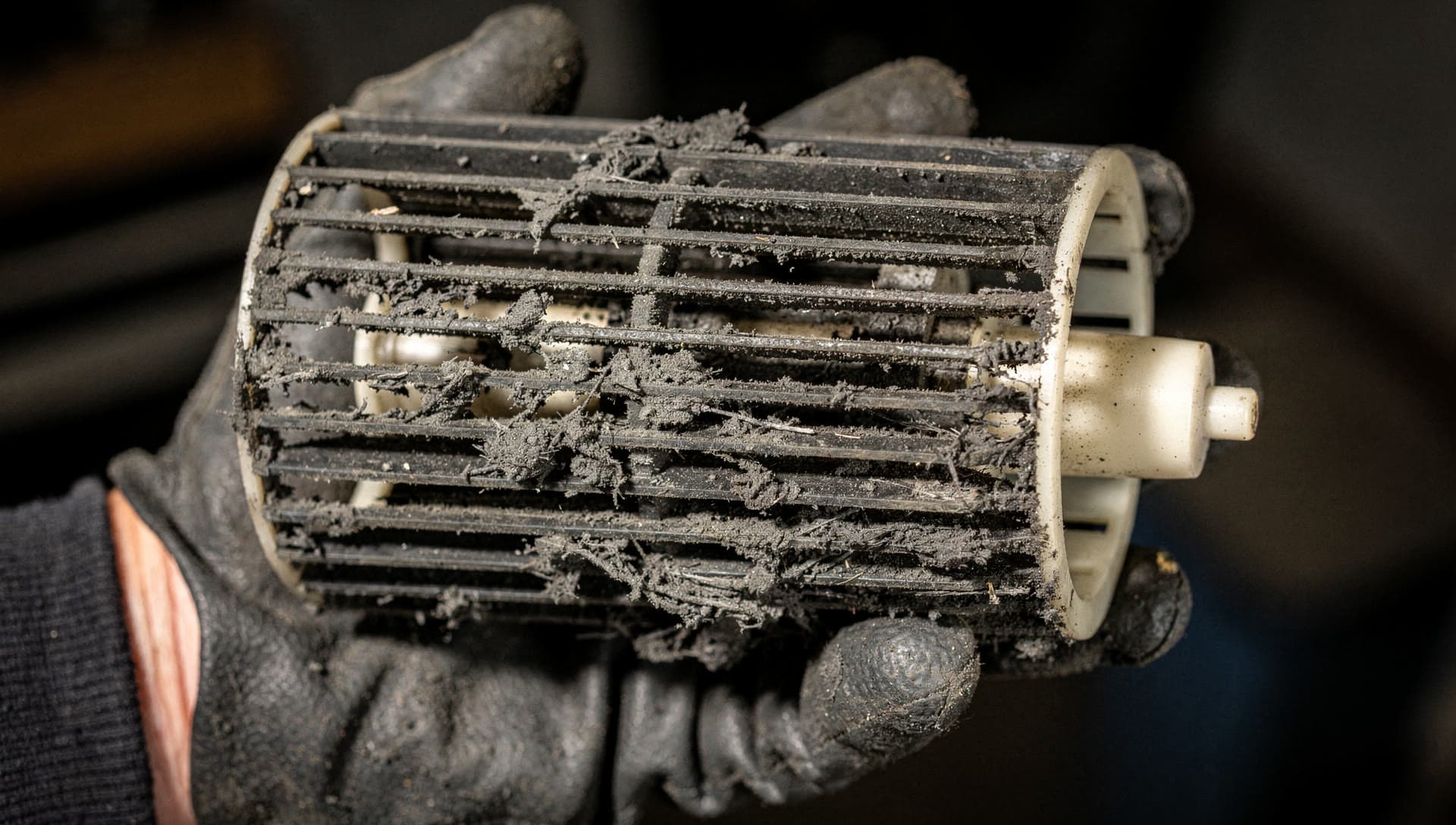 A gloved hand holding a mini split blower wheel packed solid with grey dust, straight out of the head