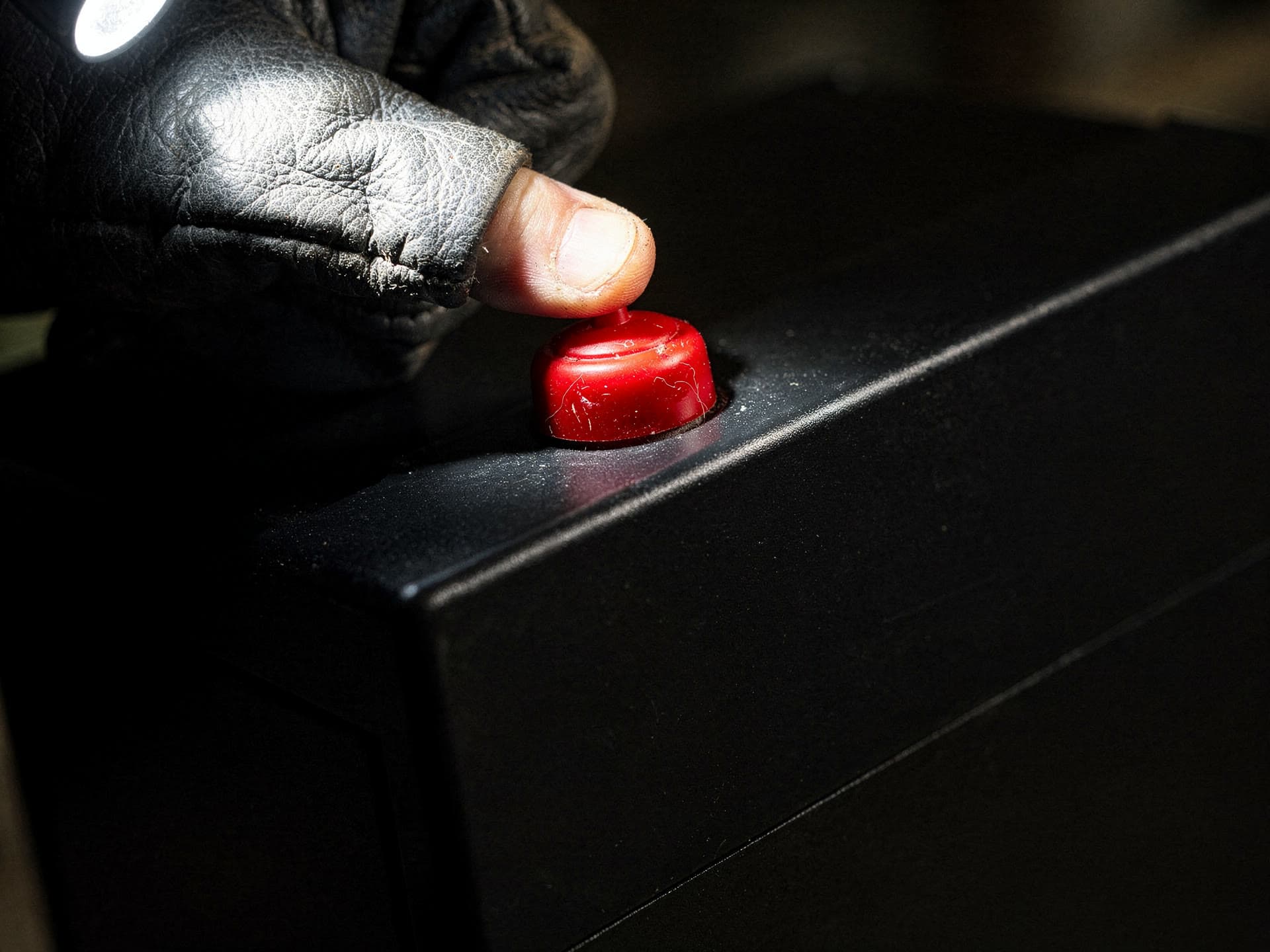 A bare fingertip pushed through the worn tip of a leather work glove to press the round red reset button on the black housing of a burner control