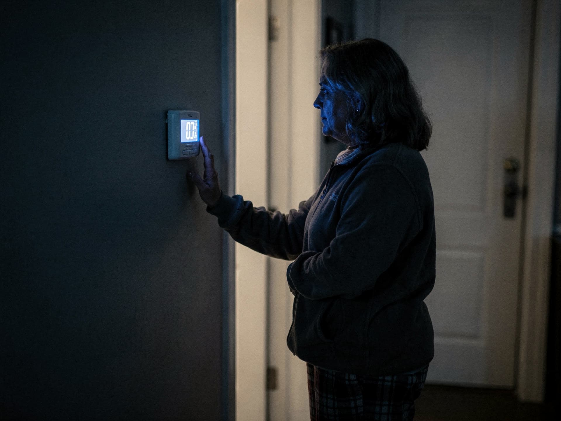 A woman in a fleece jacket and flannel pyjama bottoms standing in a dark hallway at night, pressing the button on a backlit wall thermostat, her face lit blue by the display