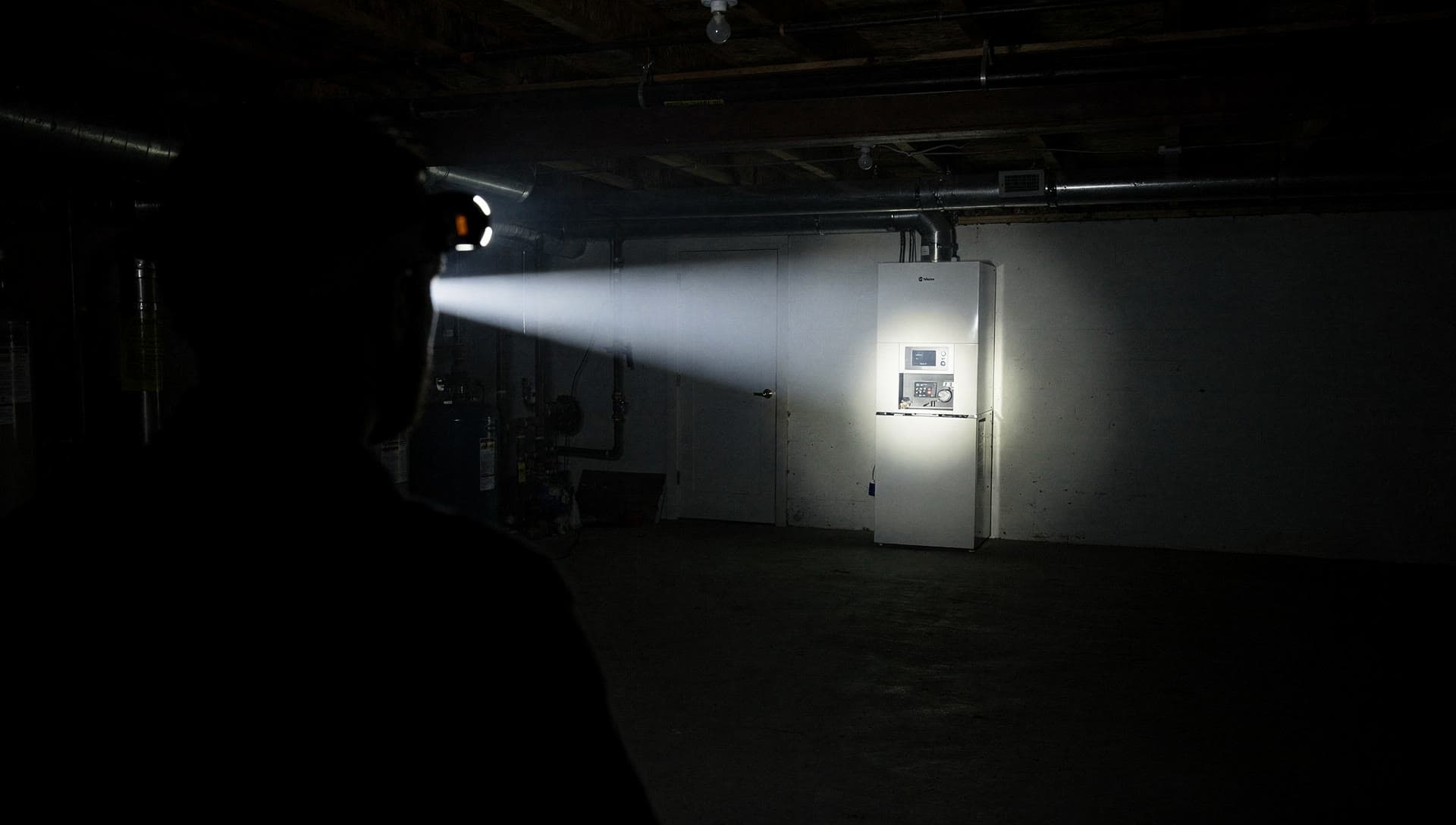 A technician in silhouette from behind in a dark basement, the beam of a headlamp cutting across the room to a tall pale heating cabinet against the far wall