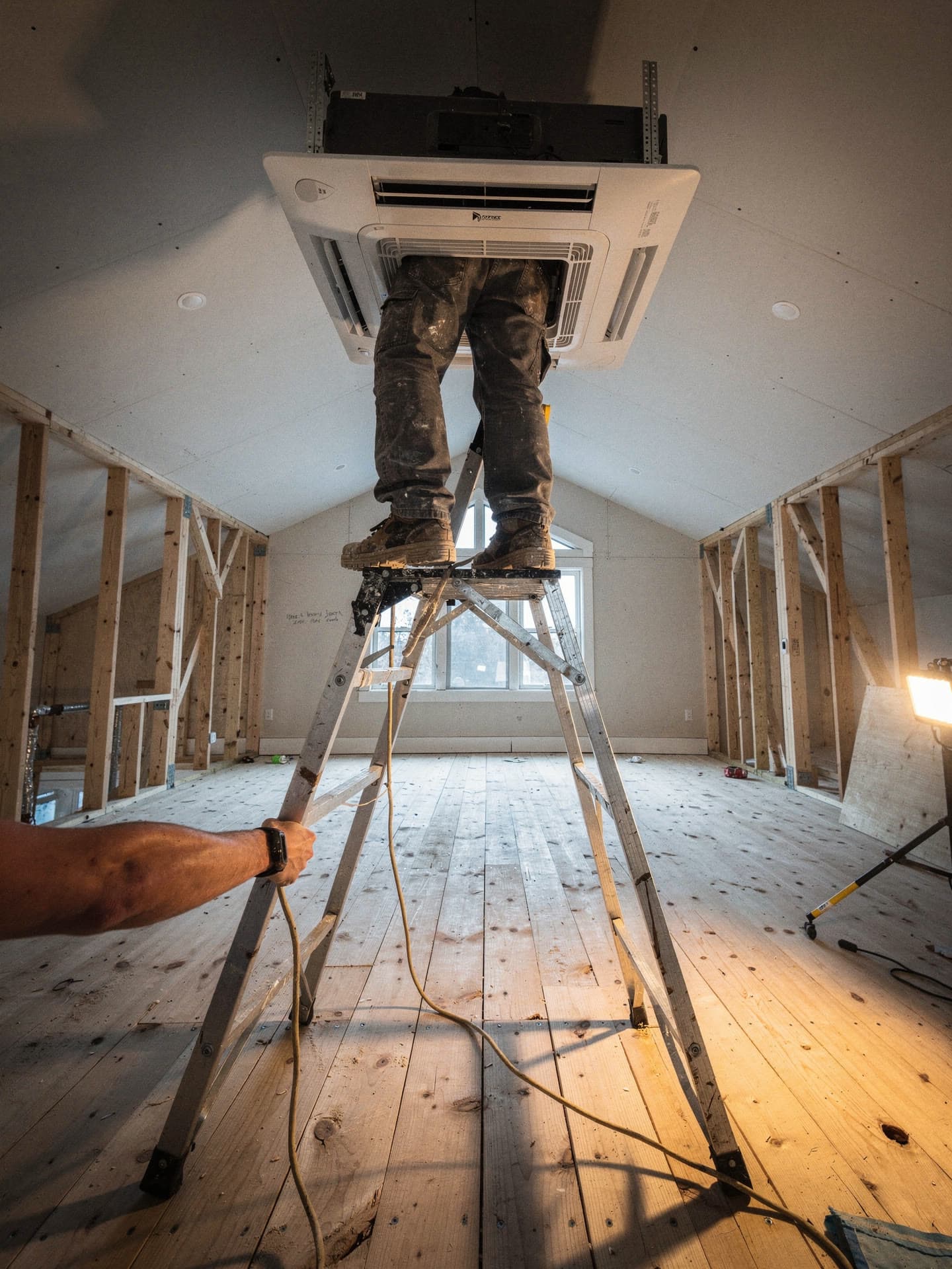 A ceiling cassette mini split head being installed from a stepladder in an unfinished framed room, a second pair of hands steadying the ladder