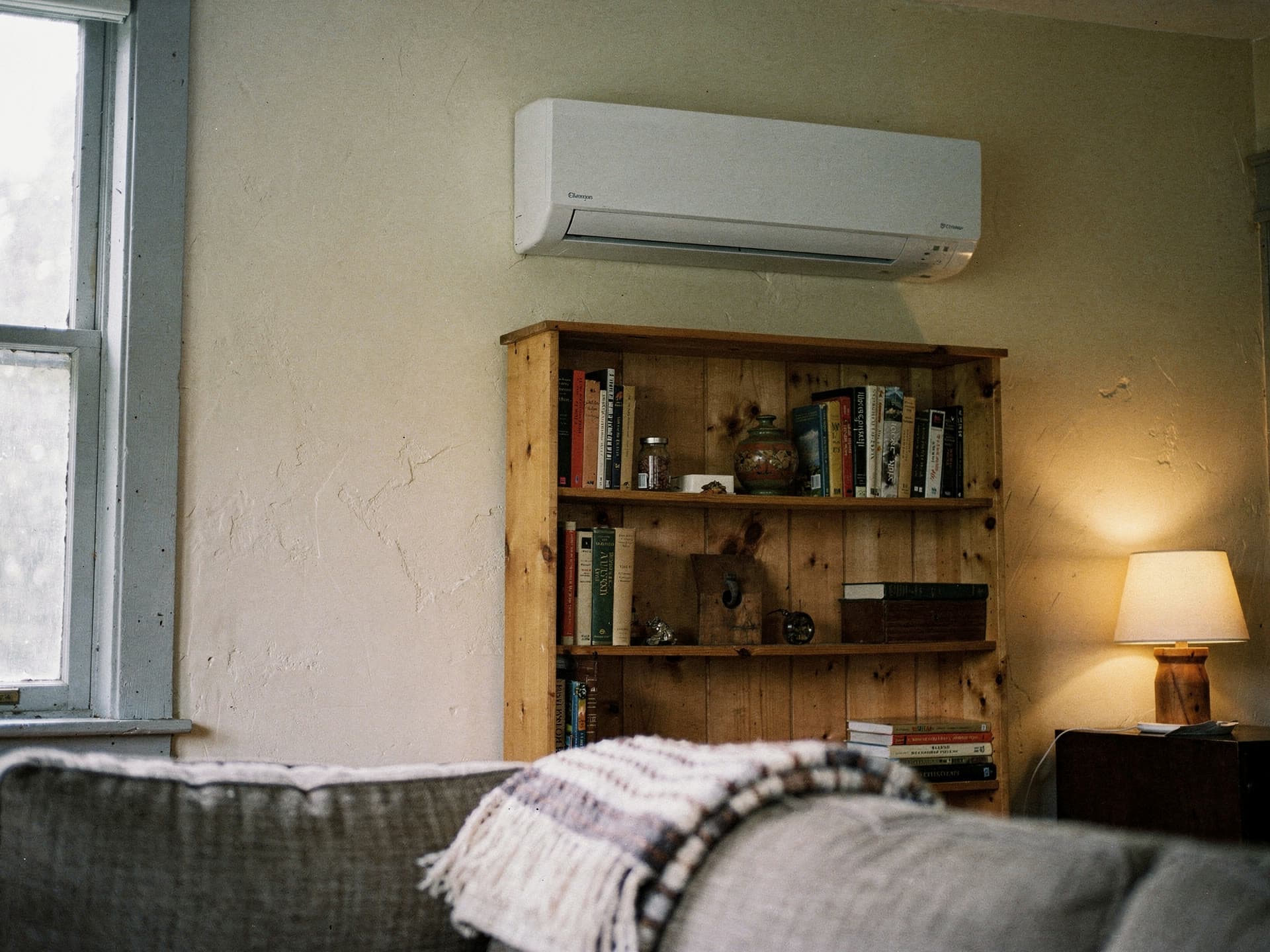 A wall mounted mini split head running high on the wall above a pine bookcase in a lived in room