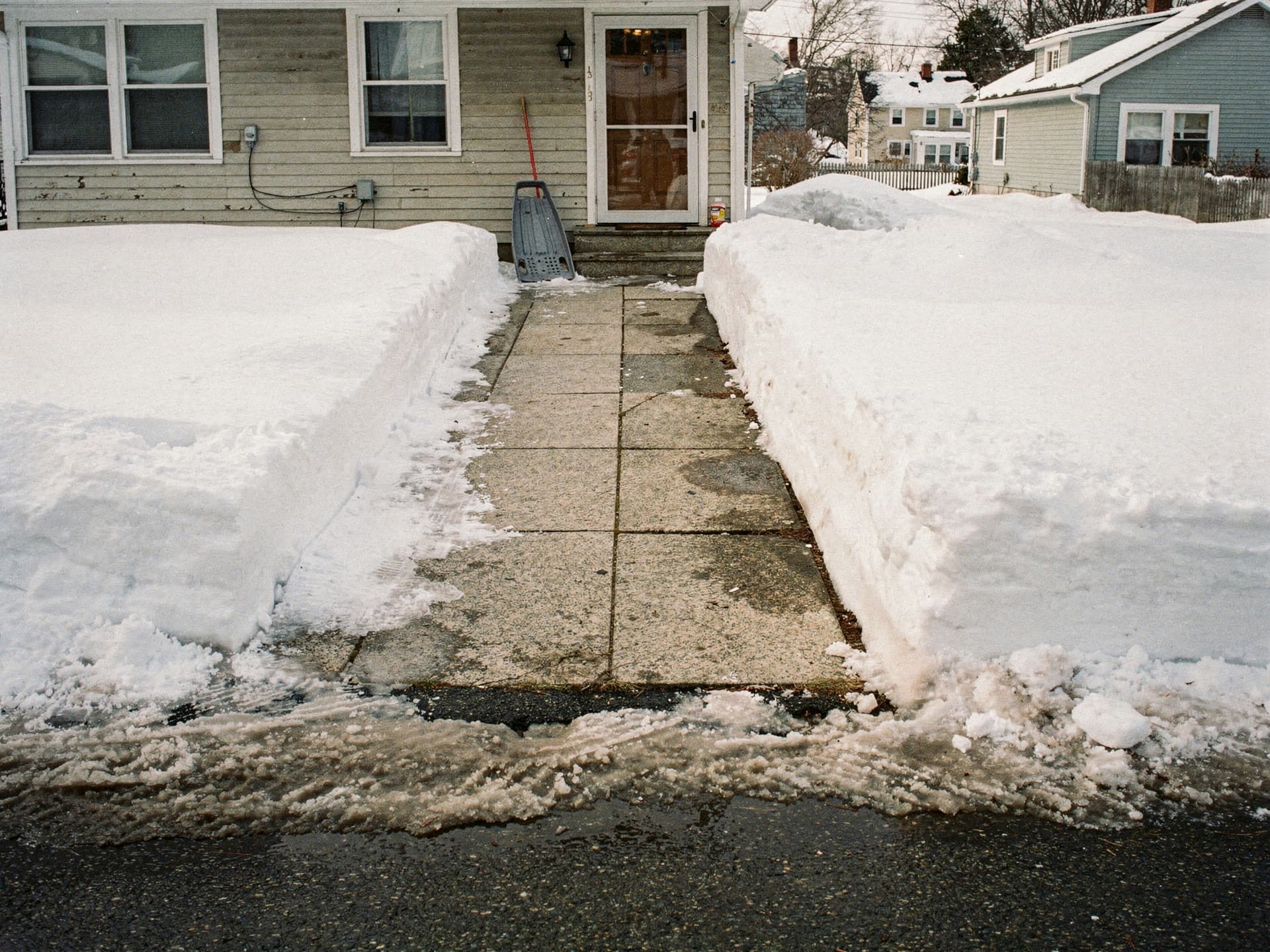A shovelled front walk cut between two snowbanks, a sled leaning by the storm door